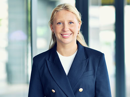 Blonde Frau lächelt, trägt dunkelblauen Blazer mit goldenen Knöpfen vor verschwommenem Hintergrund.