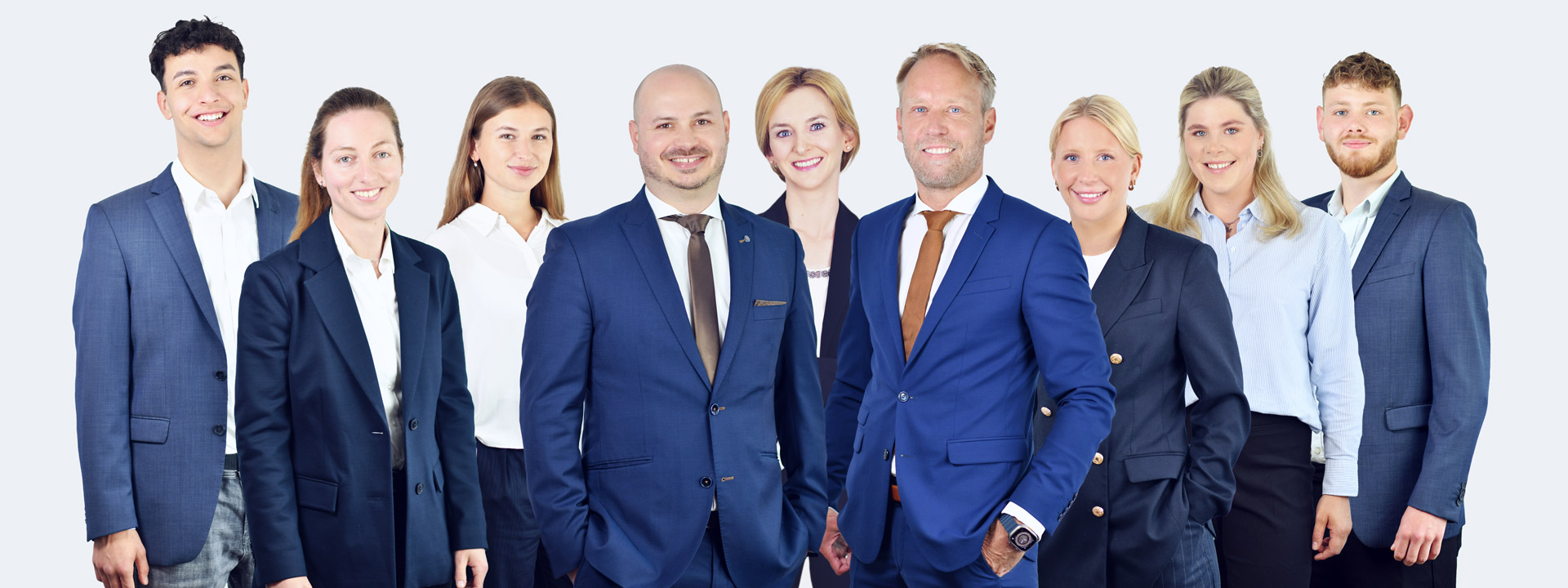 Eine Gruppe von neun lächelnden Geschäftspersonen in Anzügen vor einem hellen Hintergrund.